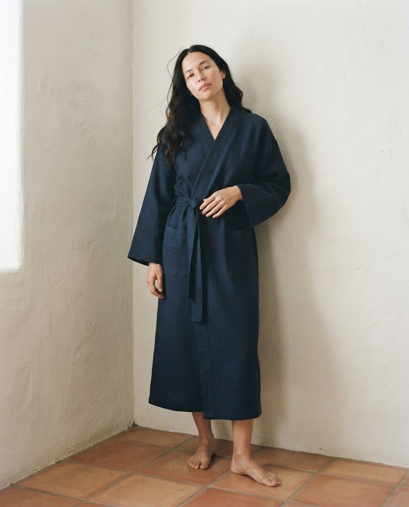 Femme en peignoir long nid d'abeille bleu marine, col V croisé et ceinture nouée, debout dans un angle de pièce chaulé