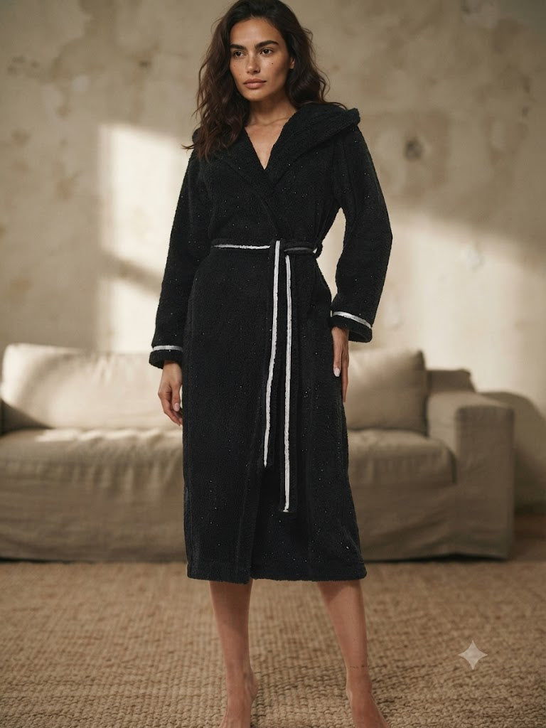 Femme debout en peignoir polaire long à capuche noir, ceinture nouée à lisere blanc, salon aux murs en chaux