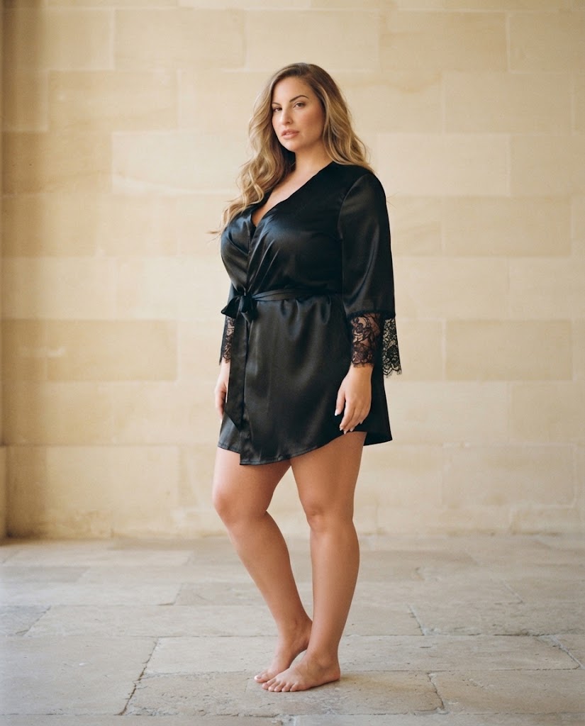 Femme en peignoir court satin noir à dentelle aux manches, debout, mur en pierre claire, peignoir grande taille femme Heure Sauvage