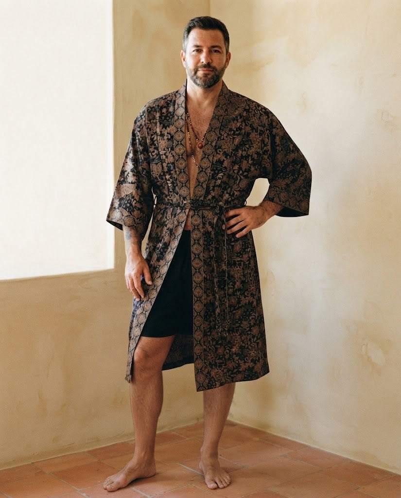 Homme debout portant un peignoir kimono long en satin noir à motifs camel, ceinture nouée, dans une pièce aux murs ocre
