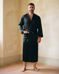 Homme debout pieds nus en robe de chambre longue en satin noir uni, col kimono et ceinture nouée, dans une pièce aux murs ocre
