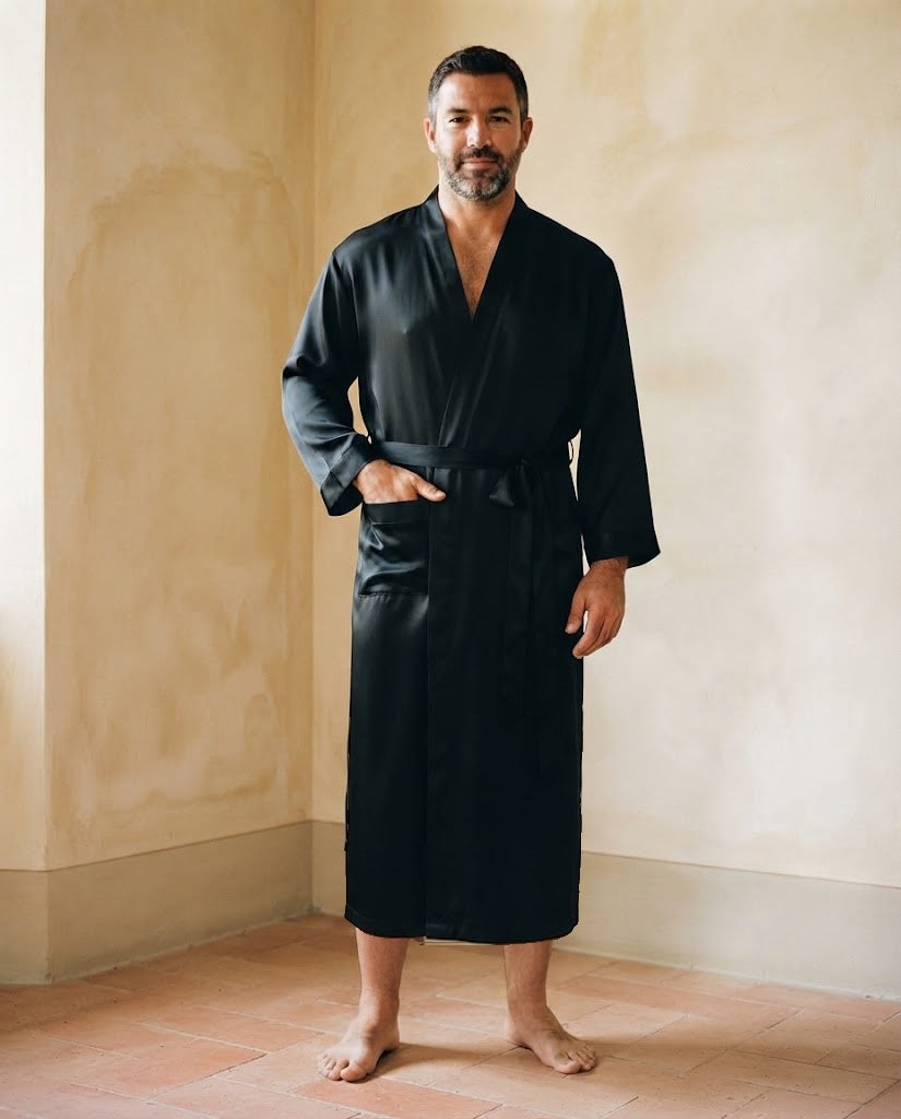 Homme debout pieds nus en robe de chambre longue en satin noir uni, col kimono et ceinture nouée, dans une pièce aux murs ocre